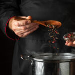 The chef adds fragrant dry peppers to a pot of boiling food. Restaurant kitchen cooking concept with advertising space on black background. Spoon in the cook hand